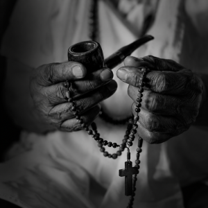 Foto em preto e branco de mãos enrugadas segurando um rosário ou cachimbo (sem exposição de entidade incorporada)